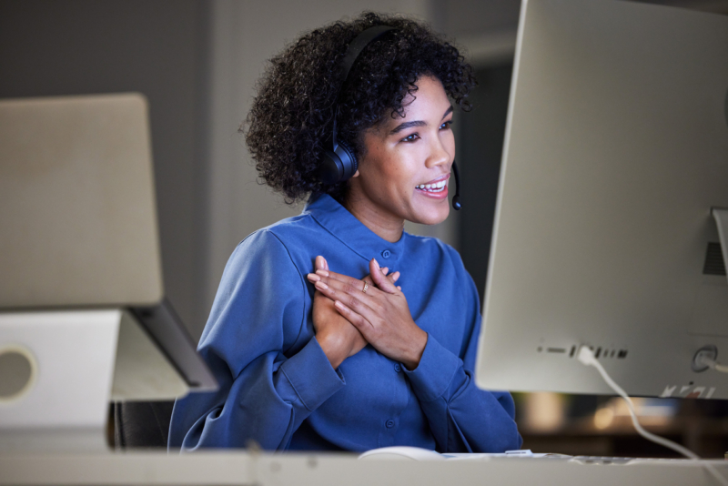 A woman working looking at a computer with a headset on, putting her hands over her heart in thankfulness. 